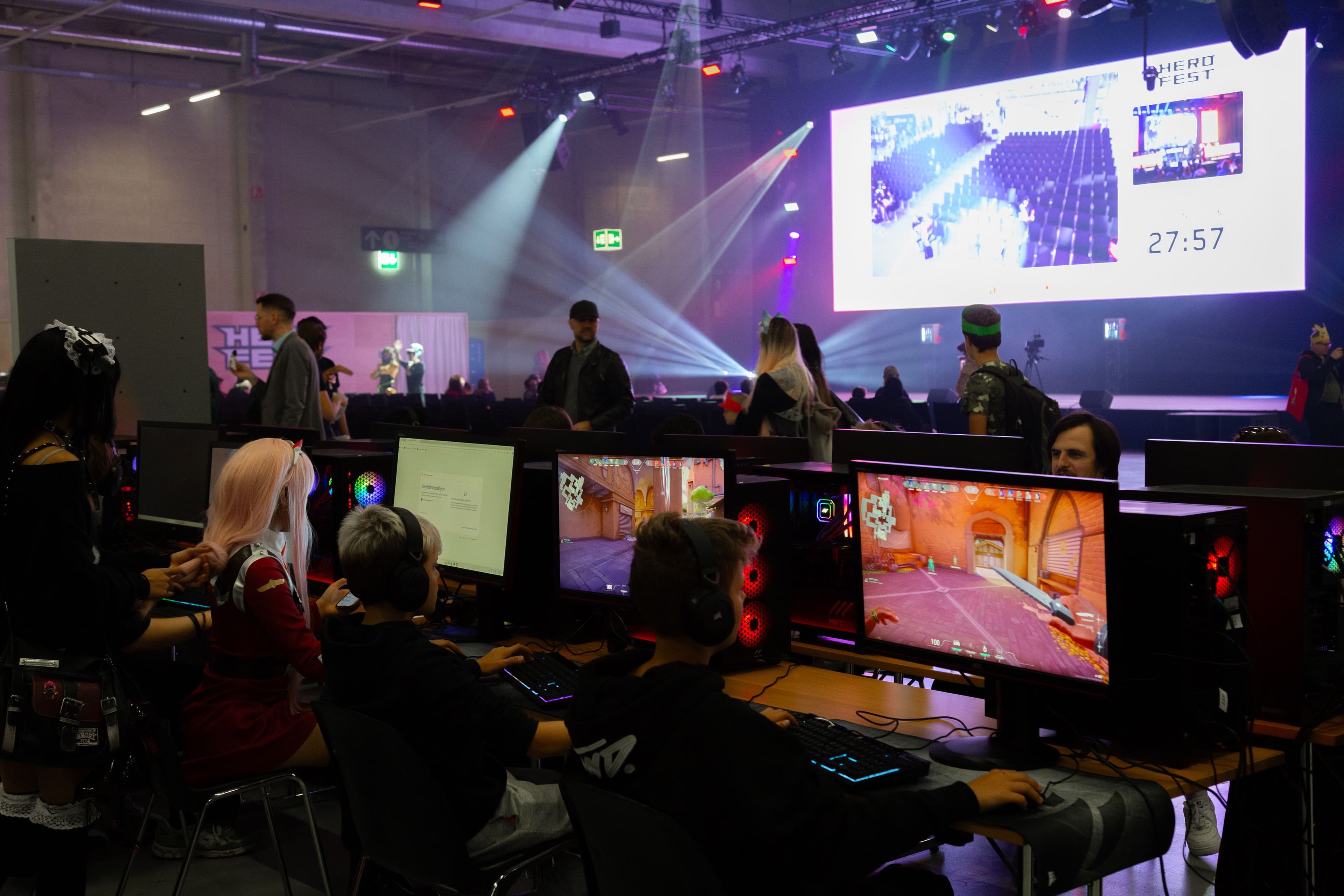 Gamer sitzen an Computern in einer Messehalle, im Hintergrund Cosplayer und eine Bildschirmwand
